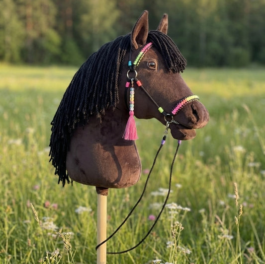 Dark brown hobby horse Ember on meadow background with yarn mane and rainbow bridle – handmade stick horse