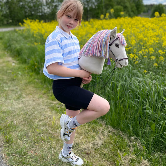 Girl riding a light grey handmade hobby horse with a colorful rainbow mane in a sunny meadow, premium stick horse toy for kids, capturing outdoor play, imagination, and hobby horsing adventures in nature