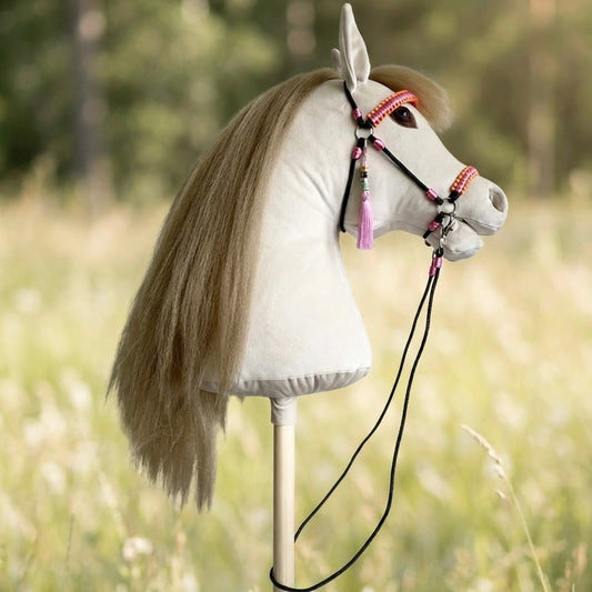 Realistic handmade hobby horse in light beige with a lifelike artificial hair mane, detailed eyes and nostrils, featuring a pink handcrafted bridle and reins with a personalized name tag and tassel, photographed outdoors in a natural summer setting