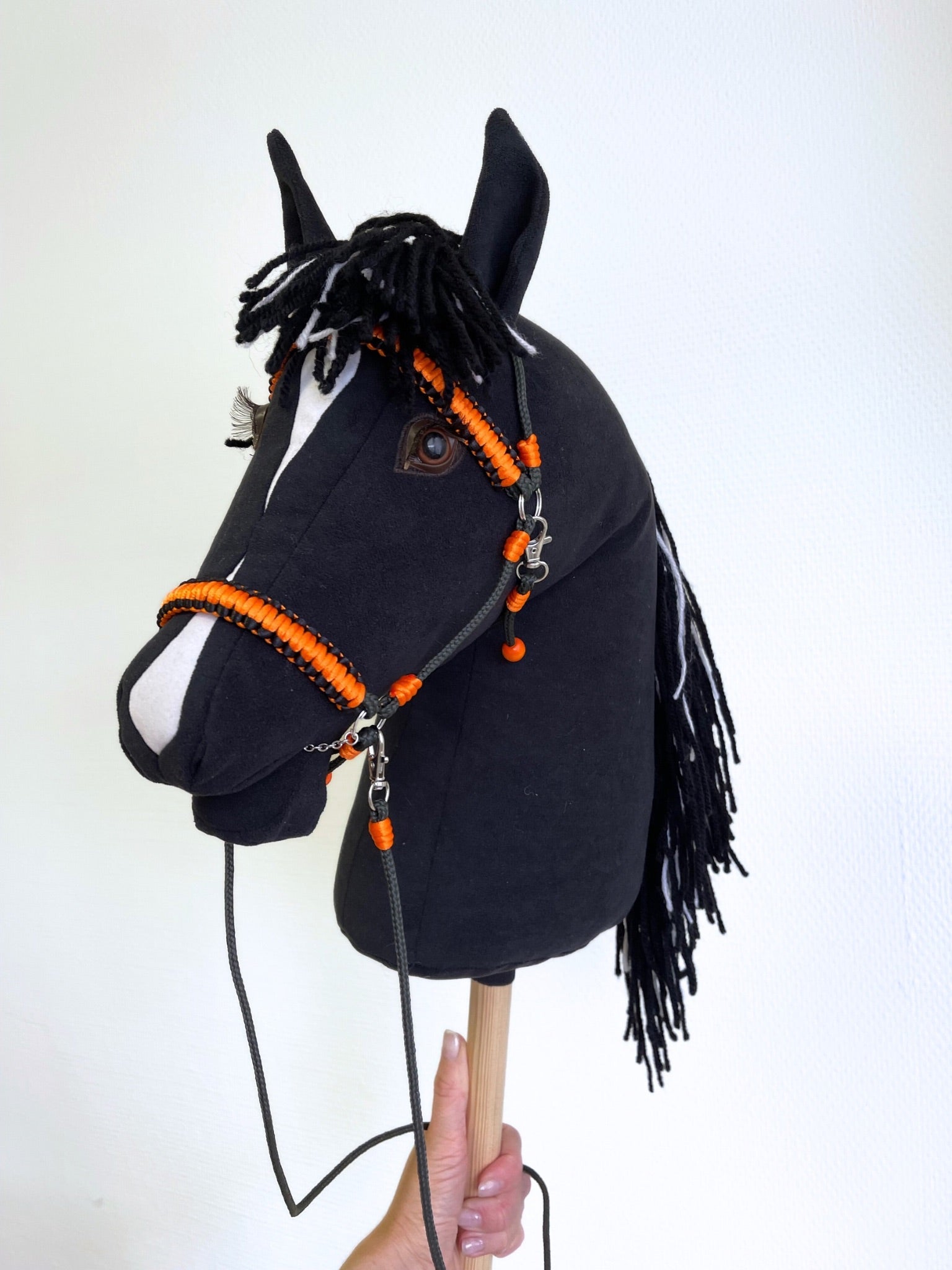 Handmade stick horse with orange bridle and name tag, standing in front of a wall display of hobby horse accessories. Unique toy horse for kids and hobby horsing fans.