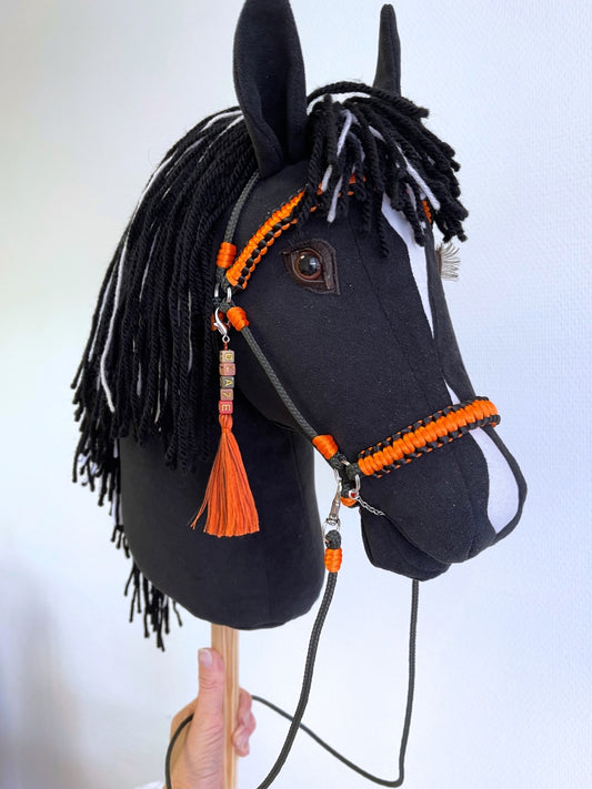 Black hobby horse with white blaze and black-and-white mane, shown with bright orange bridle, reins, and custom name charm, displayed against a neutral wall background.