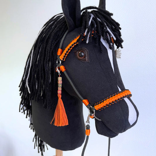 Black hobby horse with white blaze and black-and-white mane, shown with bright orange bridle, reins, and custom name charm, displayed against a neutral wall background.