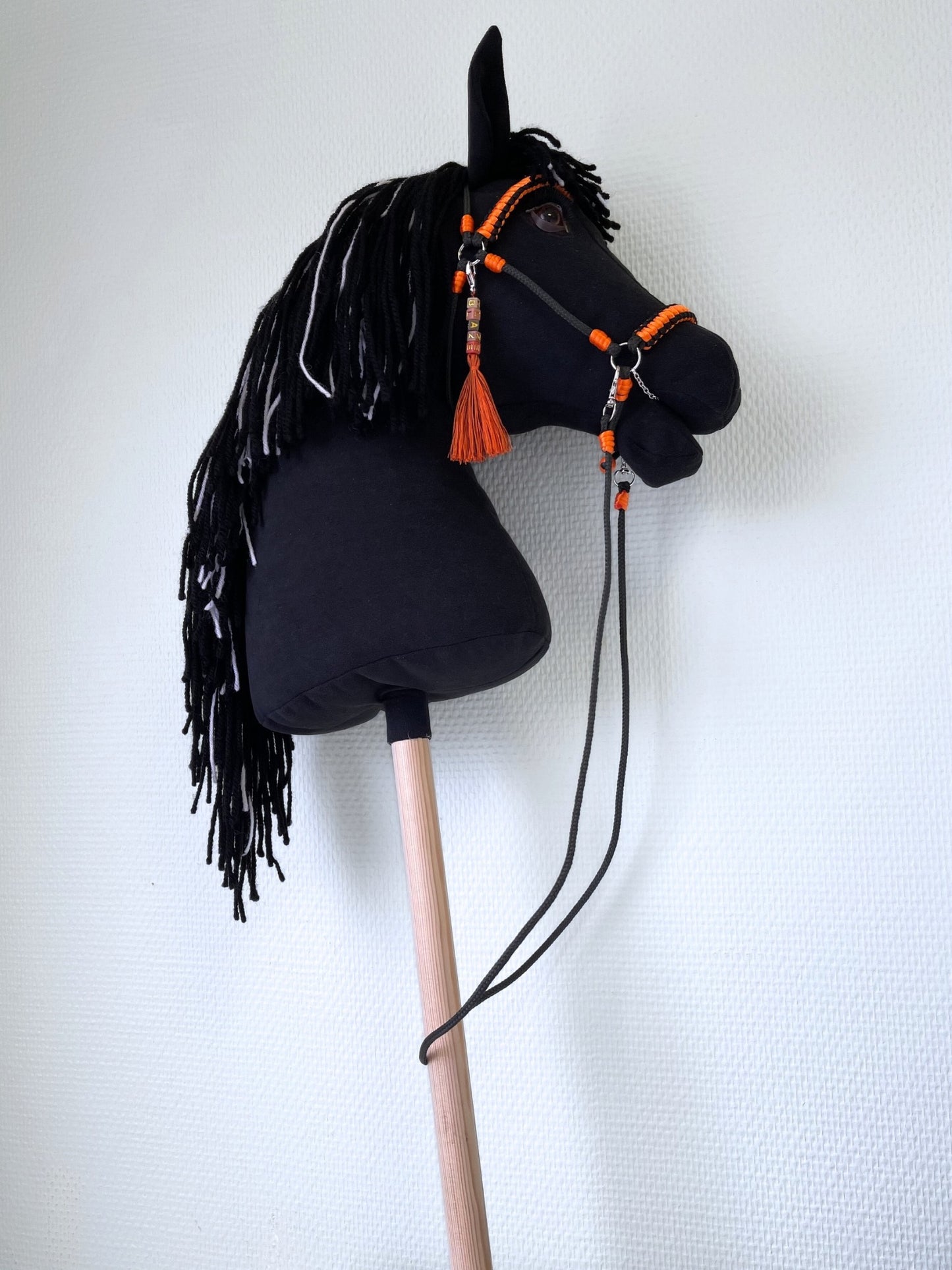 Realistic black hobby horse for hobby horsing competitions, with vivid orange bridle and reins, posed in front of an accessories display.
