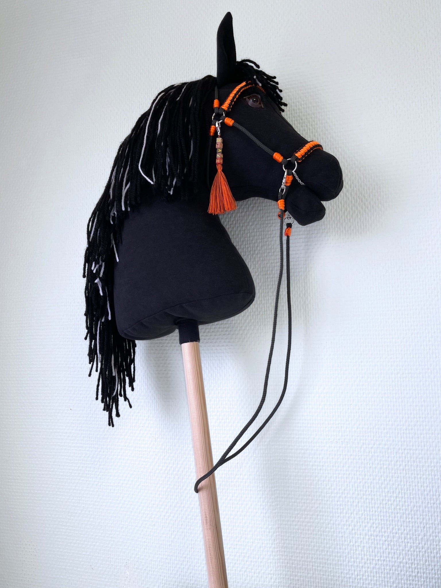 Realistic black hobby horse for hobby horsing competitions, with vivid orange bridle and reins, posed in front of an accessories display.