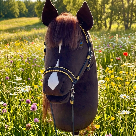 Front view of a realistic dark brown handmade hobby horse with a white blaze and lifelike artificial hair mane, featuring a dark brown bridle and reins with gold accents and a personalized tassel name tag, set against a green meadow in a natural outdoor setting