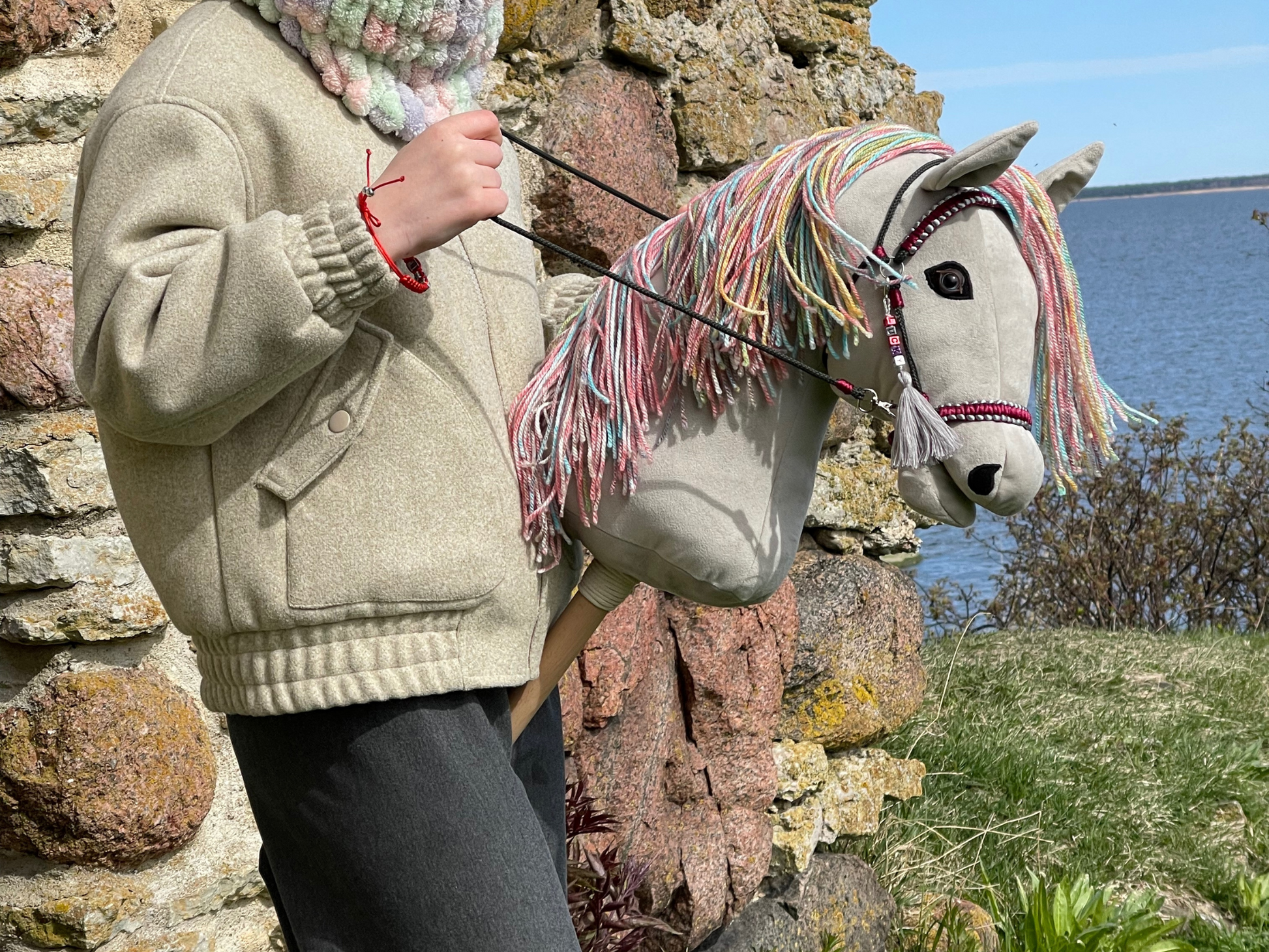 Handmade light gray hobby horse with rainbow mane and burgundy bridle, stick horse toy for hobby horsing and imaginative play.