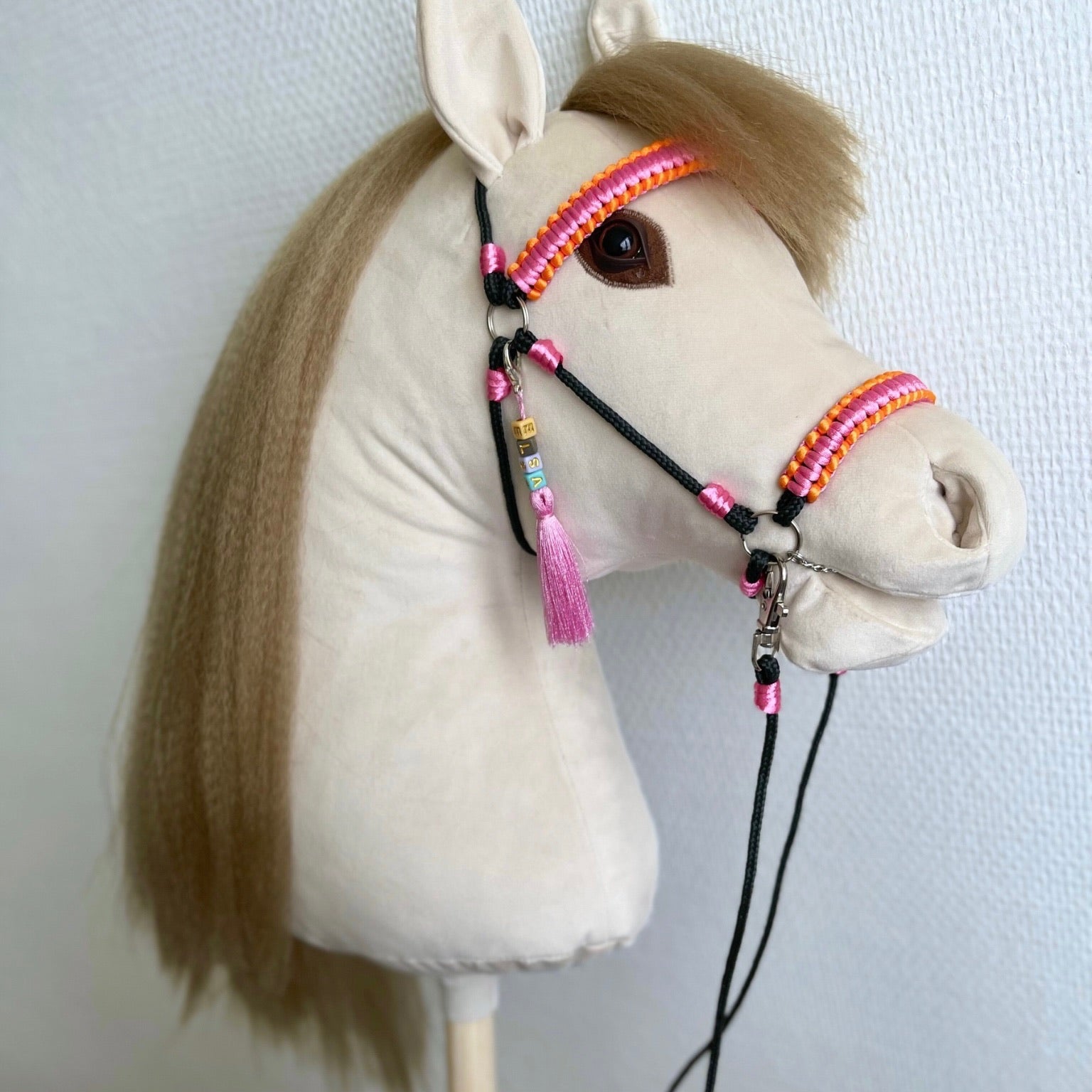 Beige handmade hobby horse on a stick with straw-colored mane, pink-orange bridle, and custom name charm.
