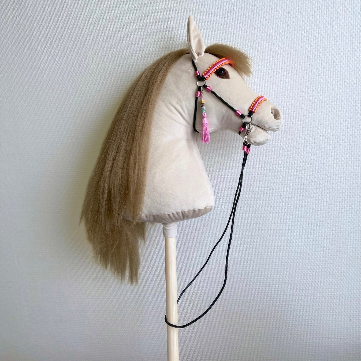 Beige handmade hobby horse with straw-colored faux hair mane, sunset pink bridle, reins, and personalized name tag.
