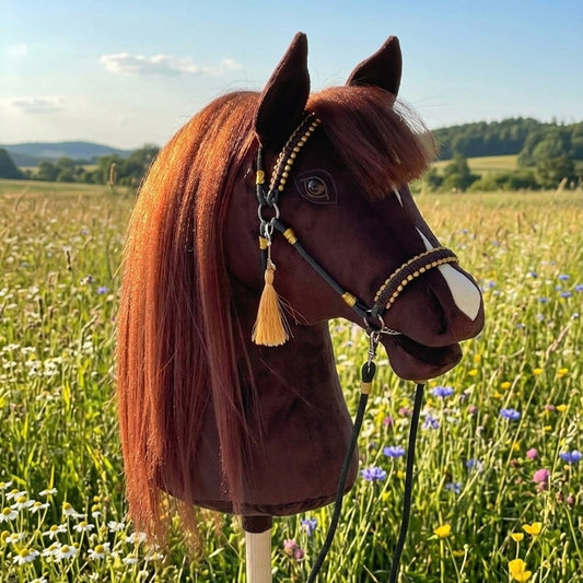 Realistic dark brown handmade hobby horse with a white blaze and lifelike artificial hair mane, featuring a dark brown bridle and reins with gold accents and a personalized tassel name tag, photographed outdoors on a green meadow in a natural setting