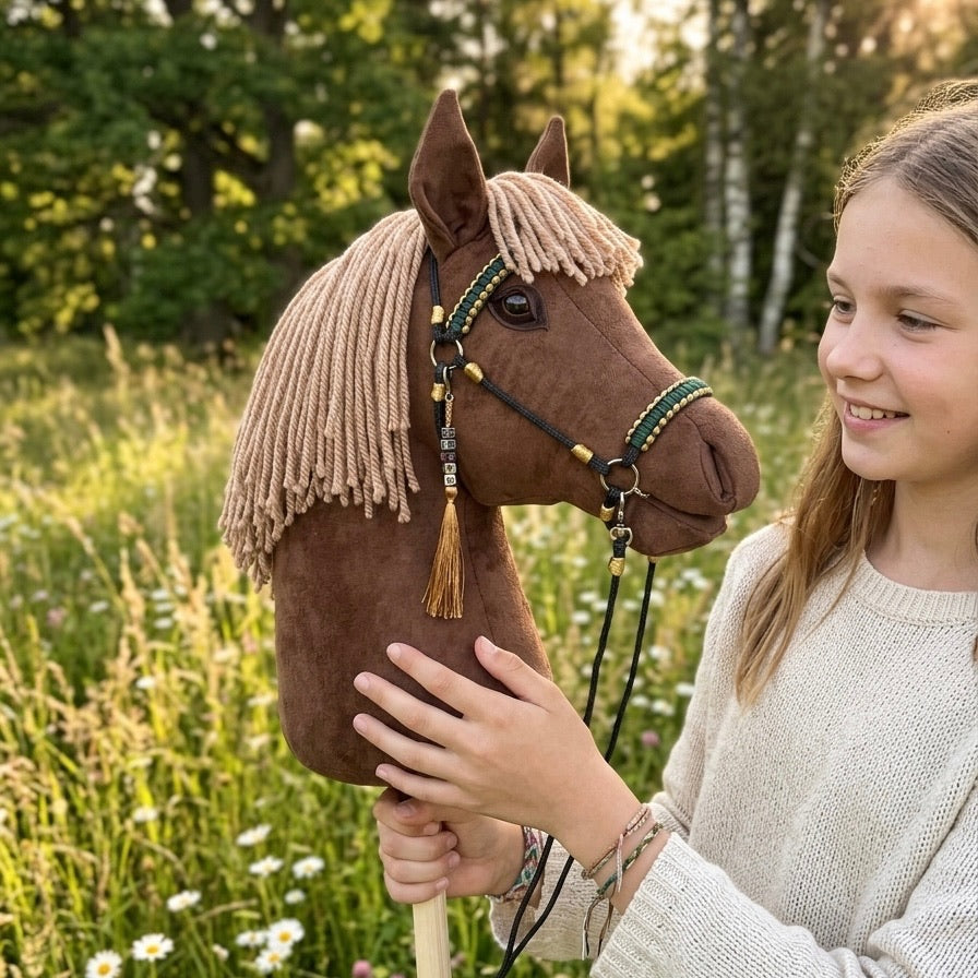 Happy girl holding a brown handmade hobby horse with light brown mane, green and gold accessories, and personalized tassel name tag.