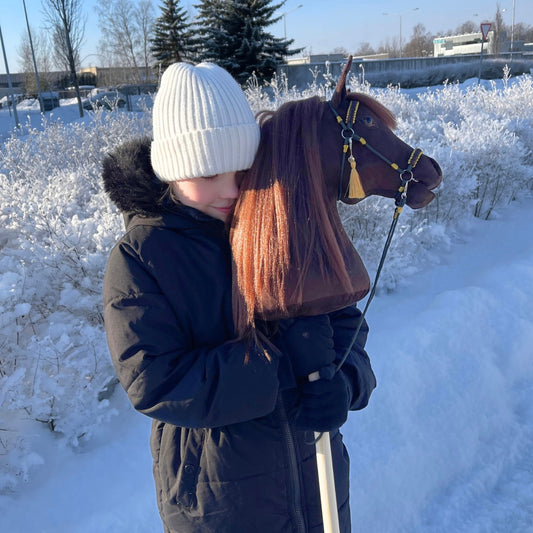 Girl holding Bella dark brown hobby horse with white blaze and golden accessories in snowy nature