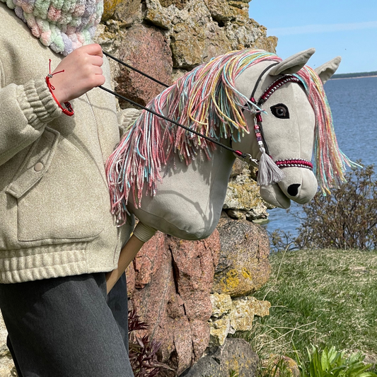 Handmade light gray hobby horse with rainbow mane and burgundy bridle, stick horse toy for hobby horsing and imaginative play.