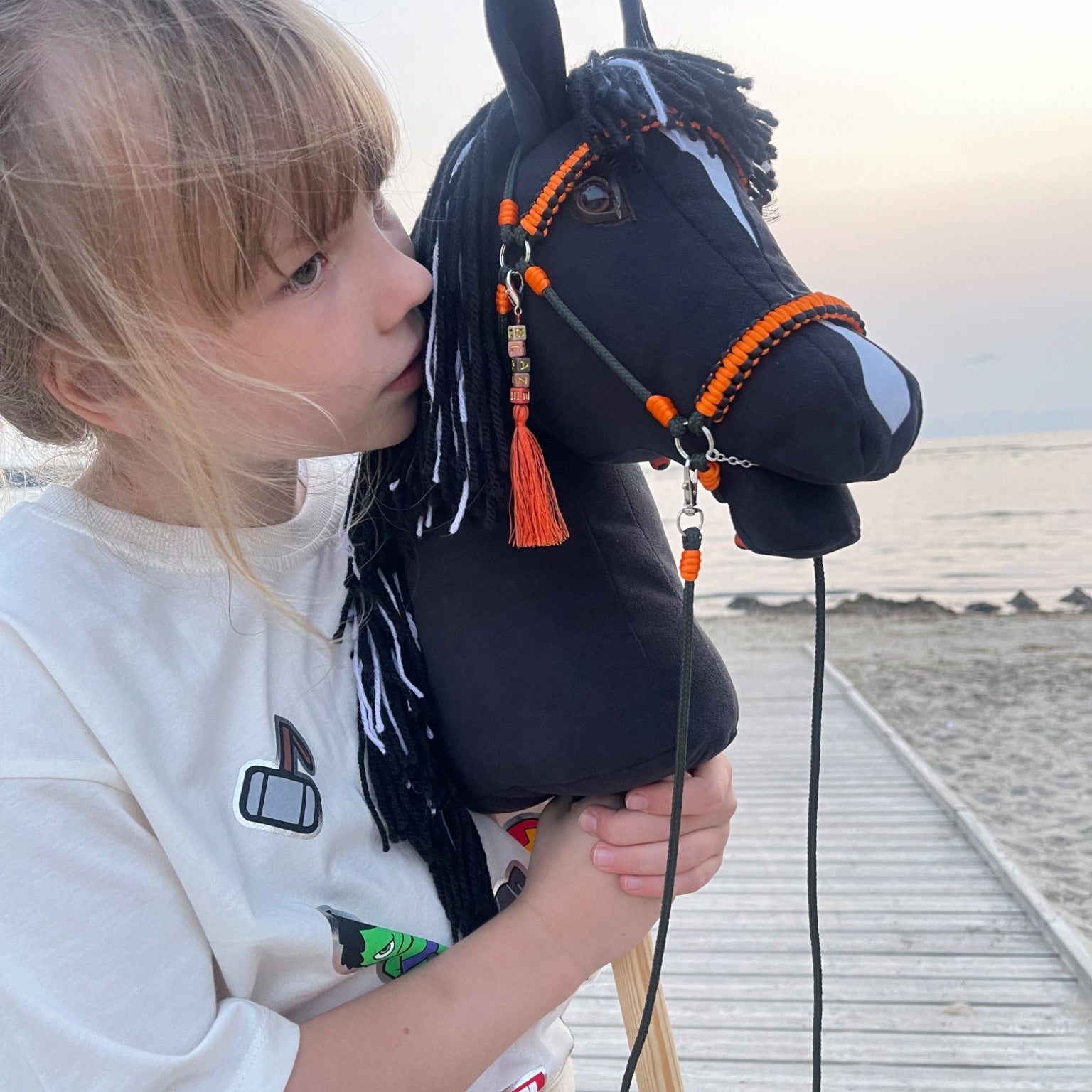 Smiling girl holding a handmade black hobby horse with white facial markings, featuring orange bridle and reins with a personalized tassel name tag, realistic details and premium craftsmanship, perfect as a custom gift for kids