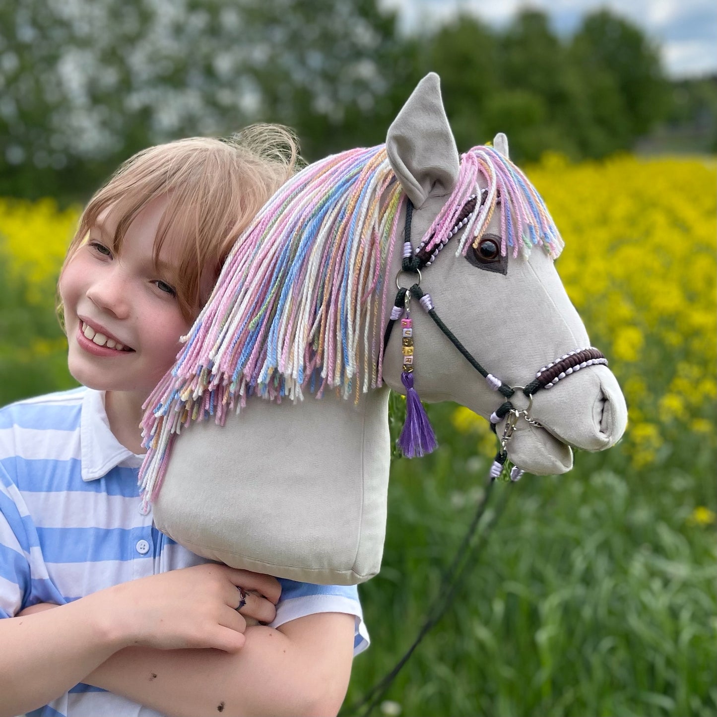 Smiling girl holding light grey handmade hobby horse with rainbow mane outdoors
